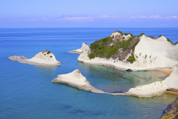 A picturesque coastal scene with striking white cliffs partially covered with greenery. The cliffs create a dramatic contrast against the clear, deep blue sea. Calm waters lap at the shore, forming small secluded bays.
