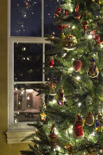 A Christmas tree adorned with various colorful ornaments is positioned near a window. The ornaments come in different shapes and sizes, including figures, fruits, and abstract designs, and emit a warm glow from the string of white lights wrapped around the tree. Outside the window, distant lights are visible, hinting at a festive evening atmosphere.