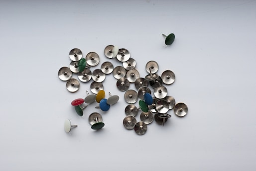 Scattered metallic push pins with several featuring colored tops in red, green, blue, white, and yellow are arranged randomly on a plain white surface.