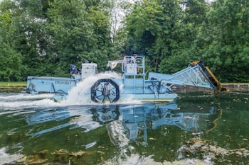 A blue aquatic machine is operating in a body of water surrounded by lush green trees. The machine has a large paddle wheel that is partially submerged, creating a splash as it moves through the water. It appears to be designed for collecting or removing debris from the water.