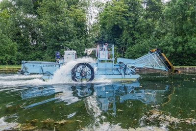 A blue aquatic machine is operating in a body of water surrounded by lush green trees. The machine has a large paddle wheel that is partially submerged, creating a splash as it moves through the water. It appears to be designed for collecting or removing debris from the water.