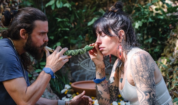 A man and a woman are engaged in a ritual or meditative practice outdoors. The man uses a pipe or instrument directed towards the woman, who holds it to her nose. Both have long hair and the woman has extensive arm tattoos. Brightly colored earrings and bracelets like wristbands are visible. The background features lush greenery and what seems to be decorative natural elements, suggesting a tranquil or ceremonial setting.