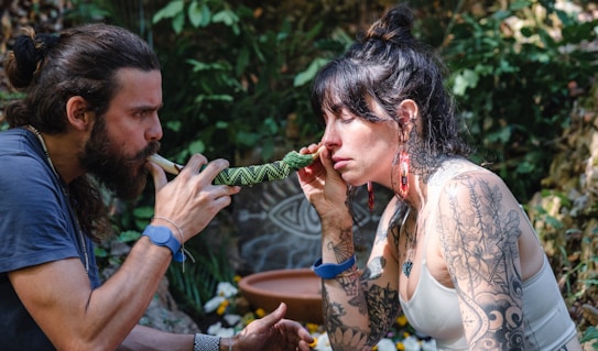 A man and a woman are engaged in a ritual or meditative practice outdoors. The man uses a pipe or instrument directed towards the woman, who holds it to her nose. Both have long hair and the woman has extensive arm tattoos. Brightly colored earrings and bracelets like wristbands are visible. The background features lush greenery and what seems to be decorative natural elements, suggesting a tranquil or ceremonial setting.