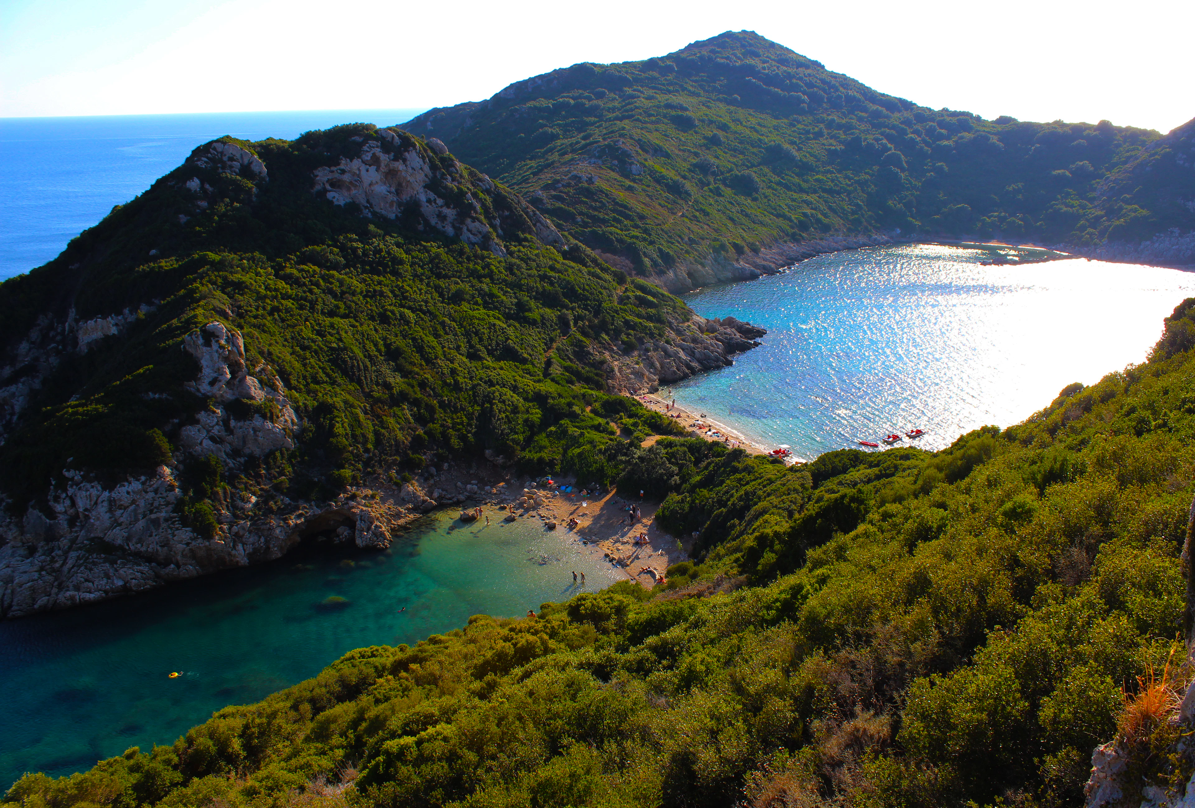 Two turquoise bays meeting on either side of a green headland on the Corfu coast, sunlight glittering on the water, seen from the scrub-covered cliffs above