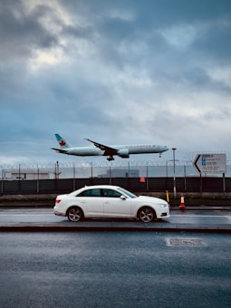 A commercial airplane from Air Canada is captured in mid-flight above a roadway. Below the aircraft, a white sedan is parked on the side of the street. The background features a fenced area with barbed wire, and a cloudy sky creates a dramatic backdrop. There are traffic cones and a direction sign visible on the sidewalk.