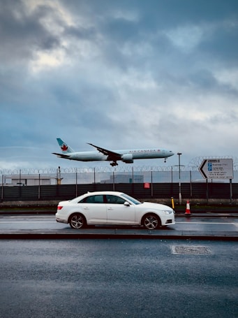 A commercial airplane from Air Canada is captured in mid-flight above a roadway. Below the aircraft, a white sedan is parked on the side of the street. The background features a fenced area with barbed wire, and a cloudy sky creates a dramatic backdrop. There are traffic cones and a direction sign visible on the sidewalk.