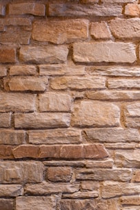 A close-up view of a textured stone wall, featuring irregularly shaped rocks that vary slightly in color. The stones are stacked horizontally, showcasing a rustic and natural appearance.