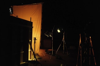 The scene appears to be staged in a dark environment with professional lighting equipment aimed at a large, orange-tinted diffusion panel or backdrop. Various tripods and stands are visible, along with some cables on the floor.