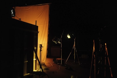 The scene appears to be staged in a dark environment with professional lighting equipment aimed at a large, orange-tinted diffusion panel or backdrop. Various tripods and stands are visible, along with some cables on the floor.