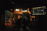 A dimly lit film studio with multiple people working amidst various equipment. Bright studio lights illuminate the set, creating a warm ambiance. A large monitor displays a scene being filmed. Posters and clothing hang on the walls, adding to the backdrop.