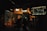 A dimly lit film studio with multiple people working amidst various equipment. Bright studio lights illuminate the set, creating a warm ambiance. A large monitor displays a scene being filmed. Posters and clothing hang on the walls, adding to the backdrop.