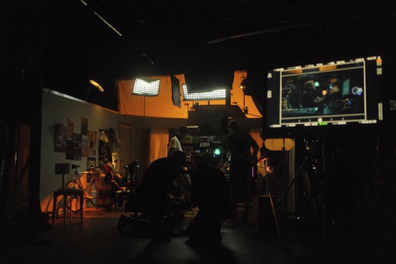 A dimly lit film studio with multiple people working amidst various equipment. Bright studio lights illuminate the set, creating a warm ambiance. A large monitor displays a scene being filmed. Posters and clothing hang on the walls, adding to the backdrop.