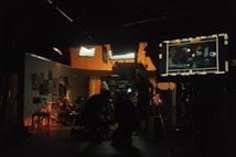A dimly lit film studio with multiple people working amidst various equipment. Bright studio lights illuminate the set, creating a warm ambiance. A large monitor displays a scene being filmed. Posters and clothing hang on the walls, adding to the backdrop.