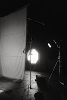 A dimly lit photography studio setup featuring a large white fabric backdrop, several lighting stands, and equipment scattered on the floor. The scene includes strong backlighting and shadows.