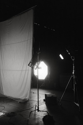 A dimly lit photography studio setup featuring a large white fabric backdrop, several lighting stands, and equipment scattered on the floor. The scene includes strong backlighting and shadows.
