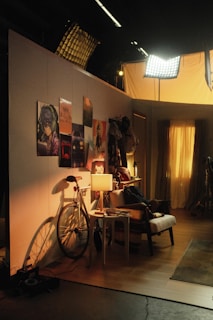 A warmly lit room with a cozy atmosphere, featuring various aesthetic elements like a bicycle leaning against the wall, several posters depicting artwork or scenes, a chair with casual items placed on it, and a small table holding books and a lamp. The setting includes curtains filtering soft light, creating a relaxed environment.