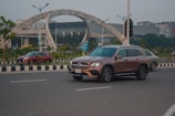 A brown luxury SUV drives along a multi-lane road with clear skies in the background. To the left, another red car and a beige sedan are visible, while in the distance, a large modern building with an arching structure and flags is situated on a landscaped area. Street lamps and trees line the road, and the setting appears urban.