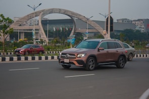 A brown luxury SUV drives along a multi-lane road with clear skies in the background. To the left, another red car and a beige sedan are visible, while in the distance, a large modern building with an arching structure and flags is situated on a landscaped area. Street lamps and trees line the road, and the setting appears urban.