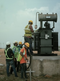 Several workers wearing safety vests and helmets are gathered around an industrial transformer. One person is elevated and appears to be working on the transformer, while others observe and assist from the ground. The setting suggests an outdoor, industrial or construction site.