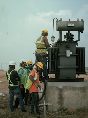 Several workers wearing safety vests and helmets are gathered around an industrial transformer. One person is elevated and appears to be working on the transformer, while others observe and assist from the ground. The setting suggests an outdoor, industrial or construction site.