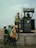 Several workers wearing safety vests and helmets are gathered around an industrial transformer. One person is elevated and appears to be working on the transformer, while others observe and assist from the ground. The setting suggests an outdoor, industrial or construction site.