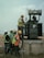 Several workers wearing safety vests and helmets are gathered around an industrial transformer. One person is elevated and appears to be working on the transformer, while others observe and assist from the ground. The setting suggests an outdoor, industrial or construction site.
