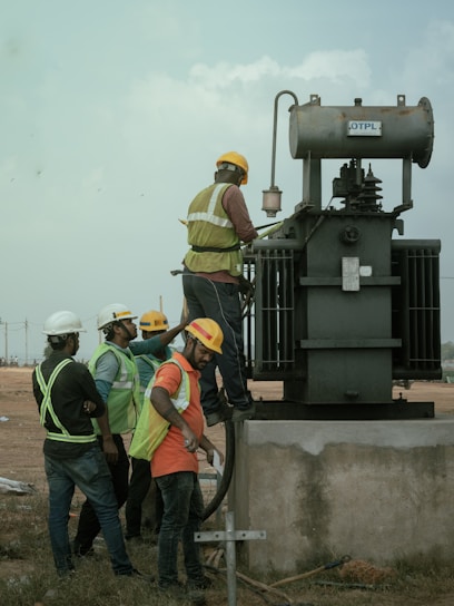 Several workers wearing safety vests and helmets are gathered around an industrial transformer. One person is elevated and appears to be working on the transformer, while others observe and assist from the ground. The setting suggests an outdoor, industrial or construction site.