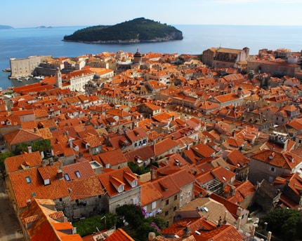 An aerial view of a historic coastal city with numerous orange terracotta rooftops. The city, situated by a calm blue sea, features old stone buildings and narrow streets. A small green island is visible in the background, surrounded by the vast ocean.