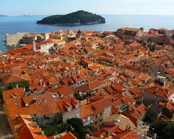An aerial view of a historic coastal city with numerous orange terracotta rooftops. The city, situated by a calm blue sea, features old stone buildings and narrow streets. A small green island is visible in the background, surrounded by the vast ocean.