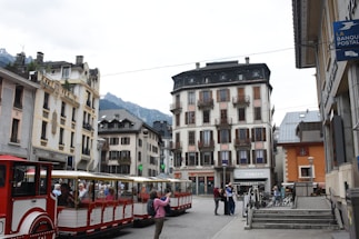 A scenic urban street view with a red and white tourist train filled with passengers in the foreground. The area is surrounded by traditional European-style buildings with balconies and shuttered windows. In the background, mountains can be seen under a cloudy sky. The scene also includes several pedestrians on the sidewalk and bicycles parked near steps.