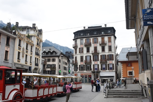 A scenic urban street view with a red and white tourist train filled with passengers in the foreground. The area is surrounded by traditional European-style buildings with balconies and shuttered windows. In the background, mountains can be seen under a cloudy sky. The scene also includes several pedestrians on the sidewalk and bicycles parked near steps.