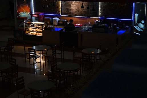A dimly lit café or coffee shop with a counter displaying pastries and a menu board behind. A person is standing behind the counter, possibly a barista, with an illuminated showcase of baked goods. Several round tables and chairs are arranged in front of the counter. Soft blue and warm lighting add an atmospheric ambiance.