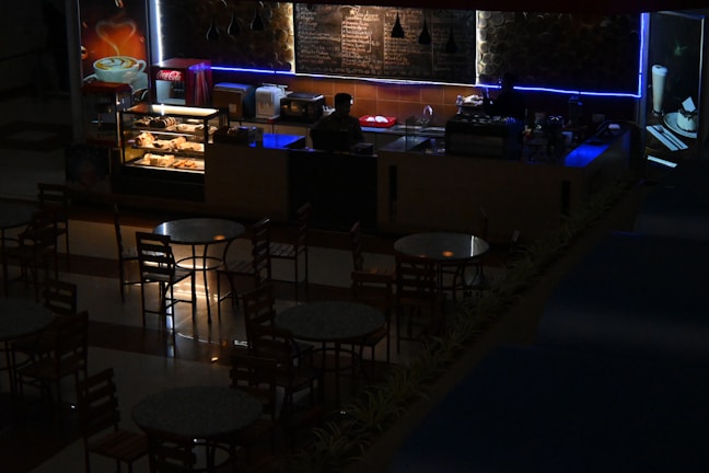 A dimly lit café or coffee shop with a counter displaying pastries and a menu board behind. A person is standing behind the counter, possibly a barista, with an illuminated showcase of baked goods. Several round tables and chairs are arranged in front of the counter. Soft blue and warm lighting add an atmospheric ambiance.