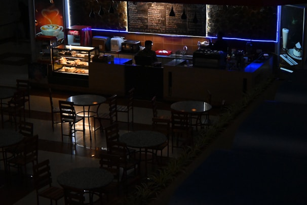 A dimly lit café or coffee shop with a counter displaying pastries and a menu board behind. A person is standing behind the counter, possibly a barista, with an illuminated showcase of baked goods. Several round tables and chairs are arranged in front of the counter. Soft blue and warm lighting add an atmospheric ambiance.
