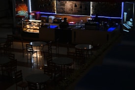 A dimly lit caf&eacute; or coffee shop with a counter displaying pastries and a menu board behind. A person is standing behind the counter, possibly a barista, with an illuminated showcase of baked goods. Several round tables and chairs are arranged in front of the counter. Soft blue and warm lighting add an atmospheric ambiance.