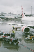 A commercial airplane is parked at an airport gate, with its fuselage and jet engine visible. Airport ground staff in yellow safety gear are handling a cart loaded with various colored suitcases on the wet tarmac. The airport terminal building is in the background, and the sky is overcast, adding a gloomy ambiance.
