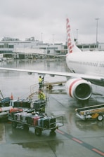 A commercial airplane is parked at an airport gate, with its fuselage and jet engine visible. Airport ground staff in yellow safety gear are handling a cart loaded with various colored suitcases on the wet tarmac. The airport terminal building is in the background, and the sky is overcast, adding a gloomy ambiance.