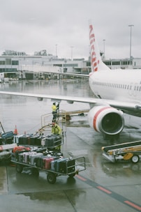 A commercial airplane is parked at an airport gate, with its fuselage and jet engine visible. Airport ground staff in yellow safety gear are handling a cart loaded with various colored suitcases on the wet tarmac. The airport terminal building is in the background, and the sky is overcast, adding a gloomy ambiance.