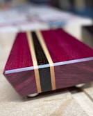 A close-up of a rectangular wooden object with a polished surface, showcasing a design with inlaid strips of different colors. The primary color is a rich purple with a central black stripe flanked by two lighter wood accents. The object is set on a smooth surface with indistinct background.