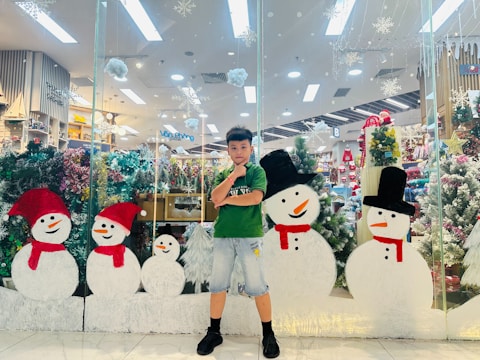 A young boy stands in front of a glass display featuring festive holiday decorations, including several snowmen with red scarves and hats. The background contains various Christmas-themed items, including decorated trees and ornaments.