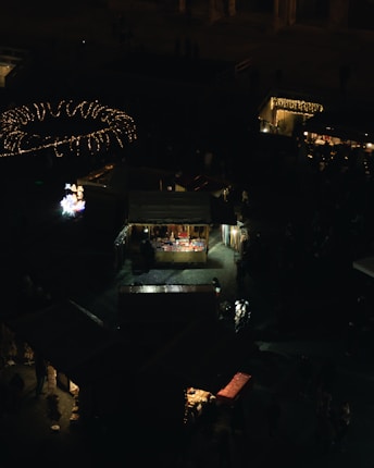 A nighttime view of a marketplace with several vendor stalls illuminated by warm yellow lights. The scene is bustling with silhouettes of people walking around. Bright decorative lights are visible above, creating a festive atmosphere.