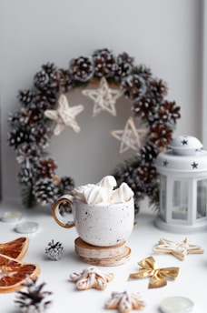 A cozy and festive scene features a white speckled mug filled with whipped cream, positioned on a wooden coaster. Surrounding the mug are decorative elements like a pinecone wreath, dried orange slices, small star-shaped decorations, and a white lantern. The setup conveys a warm, holiday atmosphere with a focus on natural and rustic accents.