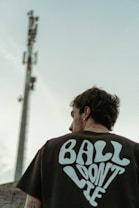 A person is seen from behind wearing a dark T-shirt with the text 'BALL DON'T LIE' printed in bold, stylized letters. The individual is standing outdoors with a large telecommunications tower visible in the background against a pale blue sky.