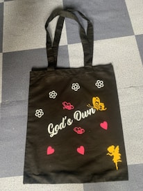 A black tote bag features white floral outlines, pink and yellow butterflies, and pink hearts. The phrase 'God's Own' is printed in white cursive lettering. A yellow silhouette of a fairy-like figure is at the bottom.