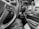 The black and white image captures the interior of a vehicle with a focus on the steering wheel. The steering wheel has control buttons for cruise control and other functionalities. A key is inserted in the ignition. The dashboard features various buttons and a gear shift, along with a phone holder. The lower portion of the image shows part of a person's leg and an open bag with documents.
