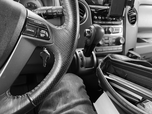 The black and white image captures the interior of a vehicle with a focus on the steering wheel. The steering wheel has control buttons for cruise control and other functionalities. A key is inserted in the ignition. The dashboard features various buttons and a gear shift, along with a phone holder. The lower portion of the image shows part of a person's leg and an open bag with documents.