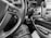 The black and white image captures the interior of a vehicle with a focus on the steering wheel. The steering wheel has control buttons for cruise control and other functionalities. A key is inserted in the ignition. The dashboard features various buttons and a gear shift, along with a phone holder. The lower portion of the image shows part of a person's leg and an open bag with documents.