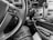 The black and white image captures the interior of a vehicle with a focus on the steering wheel. The steering wheel has control buttons for cruise control and other functionalities. A key is inserted in the ignition. The dashboard features various buttons and a gear shift, along with a phone holder. The lower portion of the image shows part of a person's leg and an open bag with documents.