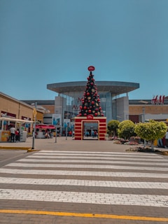 A large, decorated Christmas tree stands prominently in front of a shopping mall. The mall features modern architecture with a glass facade and visible brand signage, including H&M. People are walking and standing around the entrance. The area is well maintained with potted plants and a clear pedestrian crossing leading towards the entrance.
