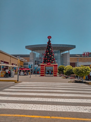 A large, decorated Christmas tree stands prominently in front of a shopping mall. The mall features modern architecture with a glass facade and visible brand signage, including H&M. People are walking and standing around the entrance. The area is well maintained with potted plants and a clear pedestrian crossing leading towards the entrance.